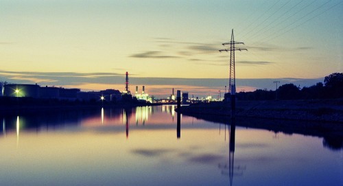 Das Bild zeigt an Industriegebiet am späten abend, es führt der Neckar vom Fotografen weg. Man sieht Schornsteine, Lichter und auf der rechten Seite eine Hochspannungs-Freileitung