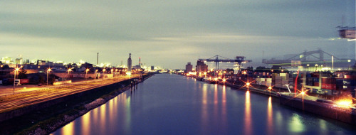 Das Bild zeigt einen Binnenhafen bei Nacht, rechts ist die Containerabfertigung, in der Mitte ein Kanal, Links Industriegebiete, es gibt viele Lichter und die Kräne haben aufgrund der langen Belichtungszeit spuren hinterlassen