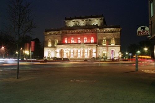 Das Bild zeigt das Theatergebäude bei Nacht, auf dem Theaterkreisel sieht man LIchtspuren der Autos