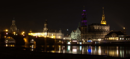 Das Bild zeigt historische Gebäude in der Altstadt von Dresden von der anderen Elbseite (Neustadt) aus. Man sieht auch den Theaterkahn und eine Elbbrücke mit Bogenmauerwerk