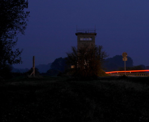 Das Bild zeigt den Beton-Grenzturm in Mattierzoll bei Nacht (von der "West-Seite"). Rechts vom Turm ist eine Straße, auf der sieht man die Rücklichter von Autos als Spuren, Links neben dem Turm ist ein Großes Baustellen-Schild, von dem man aber nur die Seite sieht.