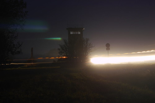 Das Bild zeigt den Beton-Grenzturm in Mattierzoll bei Nacht (von der "West-Seite"). Rechts vom Turm ist eine Straße, auf der sieht man die Schwinwerfer der Autos als Spuren, Links neben dem Turm ist ein Großes Baustellen-Schild, von dem man aber nur die Seite sieht. Von rechts nach Links durch das Bild gehen dünne Lichtspurren von einem Traktor und man sieht Reflektionen von Lichtern im Nebel