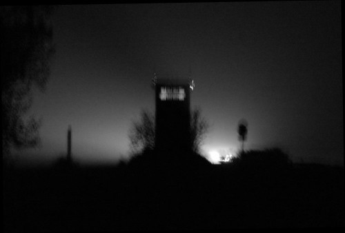 Das Bild zeigt den Beton-Grenzturm in Mattierzoll bei Nacht (von der "West-Seite"). Rechts vom Turm ist eine Straße, auf der sieht man die Scheinwerfer der Autos. Das Bild ist in schwarz/weiß und verwackelt...