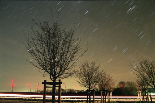 Das Bild zeigt den Nachthimmel, im Vordergrund sind ein paar Bäume und eine Straße, im Hintergrund Windkraftanlagen mit roten Positionslichtern. Die Sterne sind aufgrund der langen Belichtung Streifen