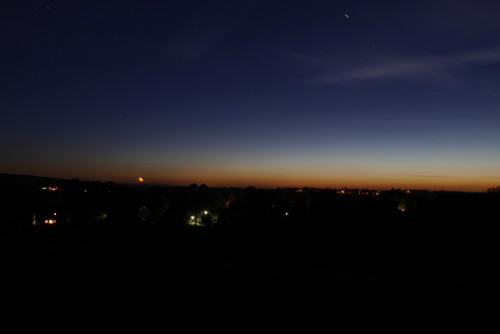 Das Foto zeigt eine Landschaft während der späten Dämmerung, man sieht ein paar Lichter an Häusern, der Himmel ist Dunkeblau und am Horizont Orange.
