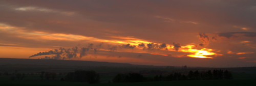 Das Bild zeigt einen Sonnenaufgang bei dichter Wolkendecke, im linken Bereich ist ein Schornstein eines Kraftwerks, der Rauch und der Rauch eines unsichtbaren Schornsteins gehen nach Rechts und sind teilweise vor der Sonne