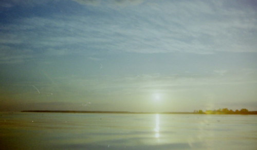 Das Bild zeigt die Sonne kurz vor Sonnenuntergang über einem ruhigen See, es ist eine Gegenlicht-Aufnahme.
