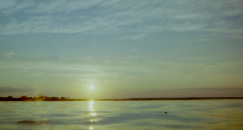 Das Bild zeigt die Sonne kurz vor Sonnenuntergang über einem ruhigen See, es ist eine Gegenlicht-Aufnahme. .