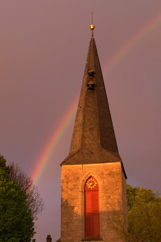 Das Bild zeigt den Kirchturm mit Uhr der Apostelkirche, hinter der Kirchturm geht ein Regenbogen durch den Himmel