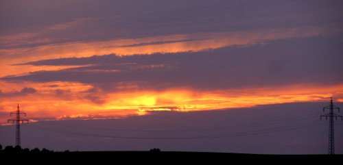 Das Bild zeigt einen Sonnenuntergang, die Sonne schaut nur durch einen kleinen Spalt zwischen den Wolken durch. Im Vordergrund sieht man ein Stück Freileitung mit zwei Masten