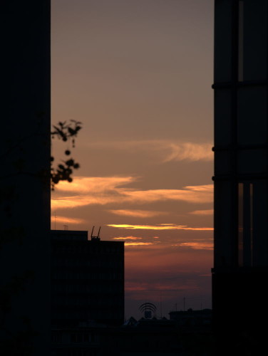 Das zeigt einen Sonnenuntergang (oder besser die Reste davon, die Sonne selbst ist nicht zu sehen, nur orange-violett leuchtende Wolken) zwischen zwei Hochhäusern