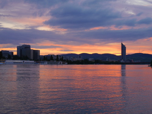 Das Bild zeigt den Sonnenuntergang an der Donau in Wien, auf dem Fluß sind Schiffe, im Hintergrund sieht man Berge und Häuser