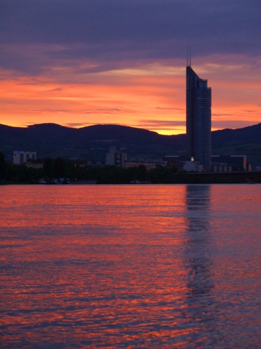 Das Bild zeigt einen Sonnenuntergang an der Donau, am Uferrand steht ein Hochhaus und weitere Häuser