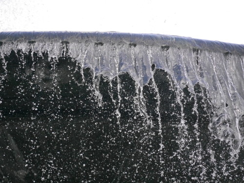 Das Foto zeigt die Kante eines Springbrunnens, über die das Wasser läuft. Aufgrund der sehr kurzen Belichtungszeit kann man einzelne Tropfen und die "Zungen" des Wassers erkennen