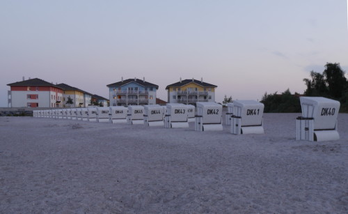 Das Bild zeigt Strandkörbe auf einem Sandstrand, im Hintergrund sieht man 3-Stöckige Hotelgebäude. Das Meer ist nicht zu sehen.
