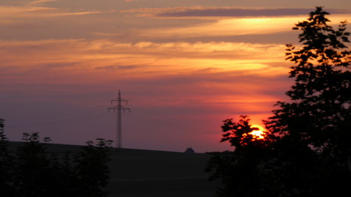 Das Bild zeigt einen Sonnenuntergang, die Sonne ist kurz vor dem Untergehen und hinter Bäumen verborgen auf der rechten Seite, im Linken drittel des Bildes steht ein Hochspannungsmast