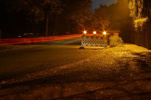 Das Bild zeigt eine Straße bei Nacht. Auf der Straße steht eine Absperrung zur Sperrung der Straße, man sieht die roten Rücklichter der Autos, die an der Absperrung vorbeigefahren sind.