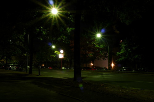 Das Bild zeigt eine nächtliche Straße in Berlin. Es ist eine Allee, rechts und LInks stehen Bäume und geparkte Atuos. Ziemlich in der Mitte steht eine Beleuchtete Uhr mit Werbetafeln.