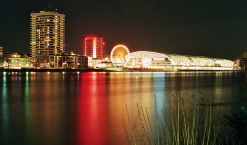 Die Nachtaufnahme zeigt ein Einkaufszentrum (die Rheingalerie), davor ein Riesenrad und beleuchtete Hochhäuser. Es spiegelt sich alles im Rhein.