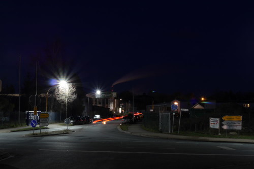Das Bild zeigt eine Straße in Schöningen, man sieht die Rücklichter eines Autos als Streifen, im Hintergrund der Schornstein vom Kraftwerk Buschhaus