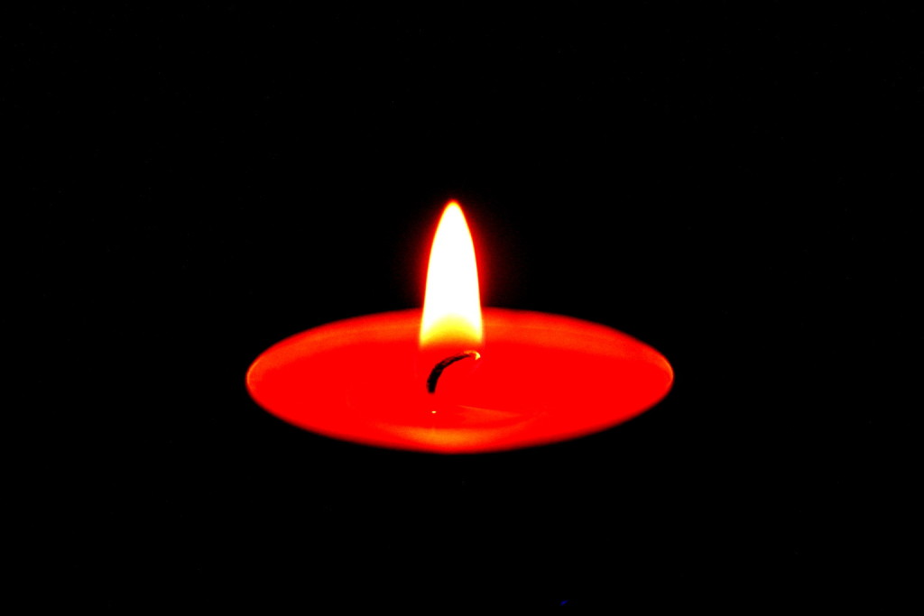 Das Bild zeigt ein brennendes, rotes Teelicht in der Dunkelheit
