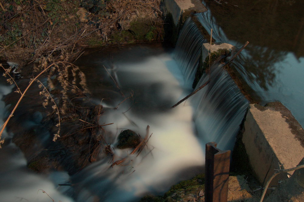 Das Foto zeigt ein Wehr in einem Bach, das Wasser strömt über das Wehr, dann ein wenig über Felsen. Im Fluss liegen Äste und ähnliches. Durch die Lange Belichtungszeit sind die Wassertropfen Silberfäden. Das BIld ist im Hochformat, man sieht viel Bach, wenig Rand.