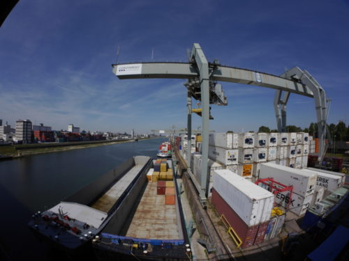 Ganz LInks sind Hafengebäude, dann das Hafenbecken, am rechten Ufer liegen zwei Schiffe, daneben der Containerkran und Containerstapel. Durch das Fischaugen-Objektiv ist das Bild rund verzerrt..