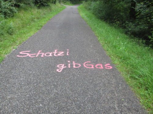 Das Foto zeigt einen geteerten Waldweg. Auf den Weg hat jemand den Satz "Schatzi gib Gas" mit pink-Neonfarbe gesprüht/gemalt