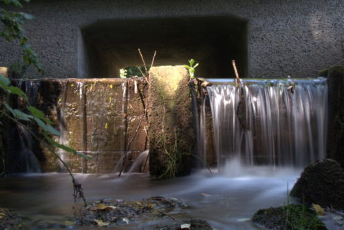Das Foto zeigt ein hölzernes Wehr in einem Bach, über das Wehr strömt das Wasser und es kommt zu einem Wasserfall. Durch die lange Belichtungszeit sieht man das Fliessen.