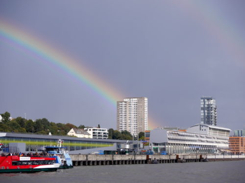 Das Foto zeigt einen Regenbogen im Hamburger Hafen, das Bild ist von der Elbe her aufgenommen und ein Hochhaus schneidet durch den Regenbogen.