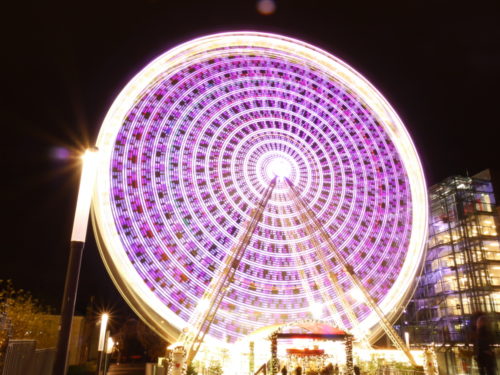 Ein Farbfoto von einem Riesenrad. Durch die Drehbewegung sind die einzelnen Lichter als Lichtspuren dargestellt.