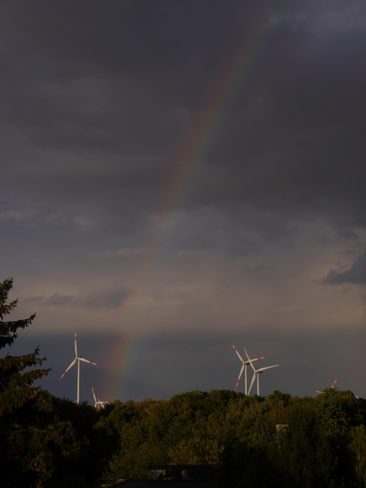 Das Foto zeigt einen Regenbogen am dunkelen Himmel, der Regenbogen beginnt am Boden bei Windkrafträdern