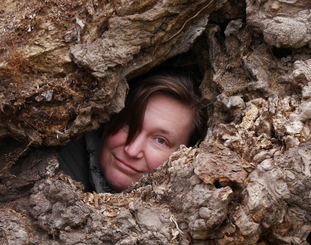 Das Foto zeigt das Gesicht einer Frau, die durch ein Loch in der Wurzel eines umgestürtzen Baums nach außen in Richtung Fotograf blickt