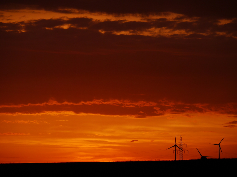 Das Foto zeigt eine Landschaft beim Sonnenuntergang, der Himmel ist Orange gefärbt. Im rechten drittel des Bildes sieht man drei Windkraftanlagen und einen Freileitungsmast.