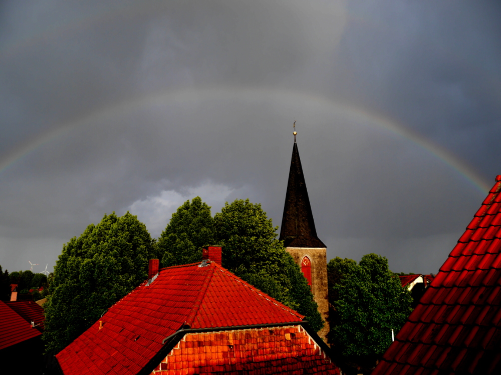 Die Landschaftsaufnahme zeigt einen Regenbogen über den Dächern und über dem Kirchturm etwa in Bildmitte.