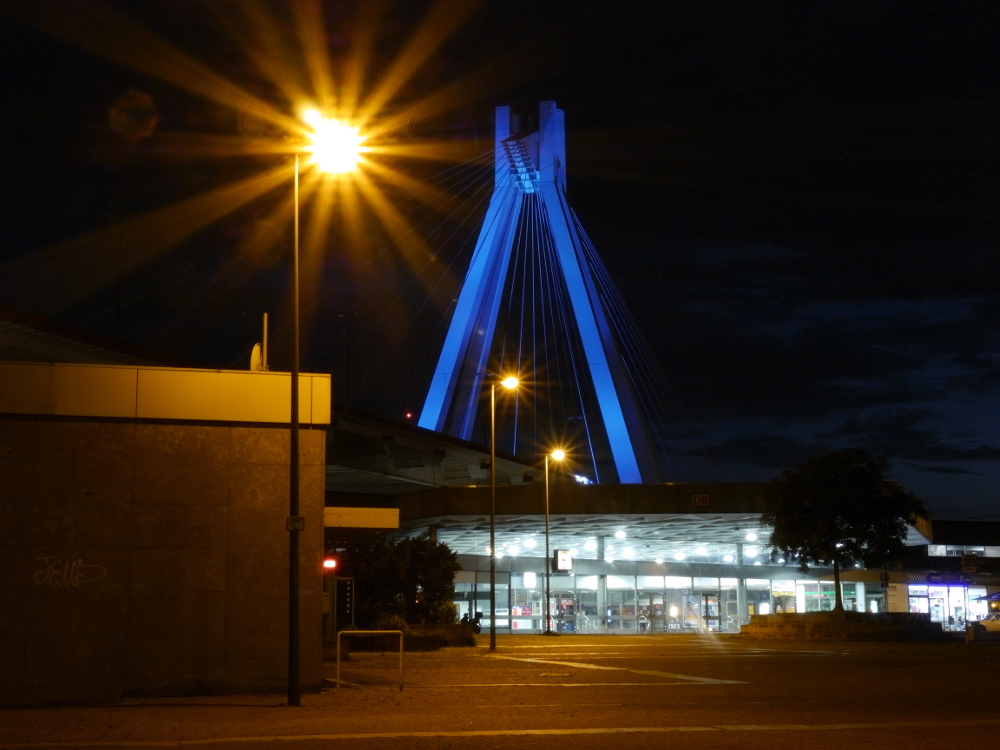 Das Bild zeigt den Hauptbahnhof von Ludwigshafen. Gut sieht man die Brücke, die sich über den Hauptbahnhof spannt. Es ist eine Nachtaufnahme, die Brücke wird blau angestrahlt.