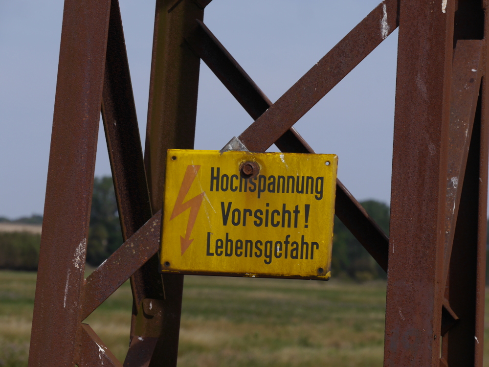 Ein gelbes, schon angerosteten Warnschild "Hochspannung, Vorsicht! Lebensgefahr" an einem verrosteten Stahlgittermast.