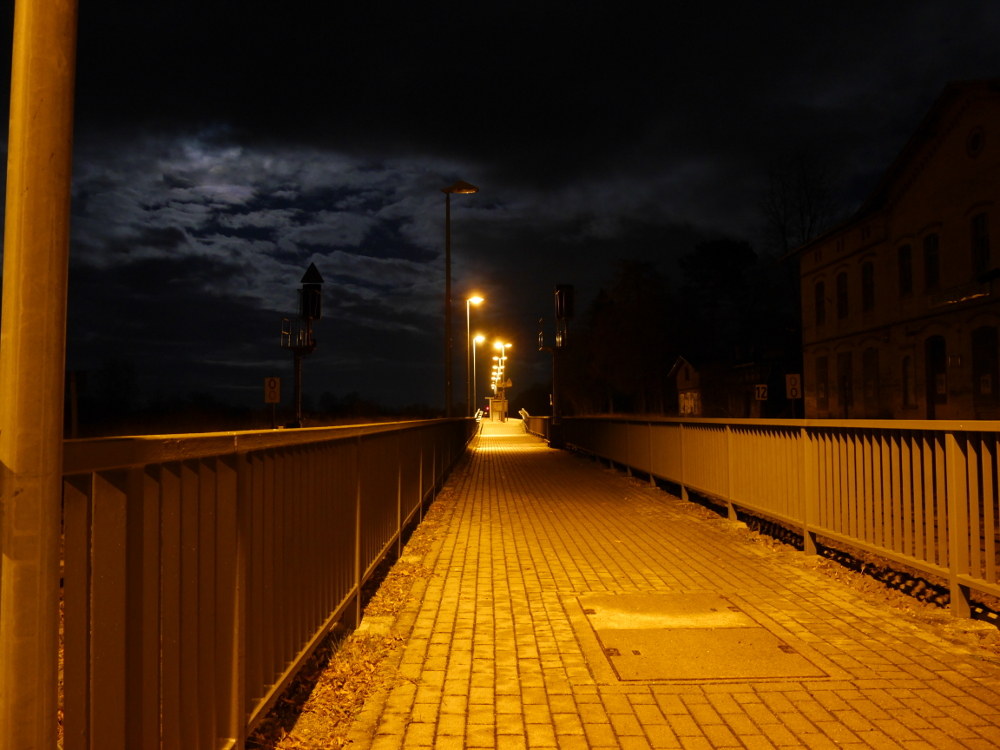 Die Nachtaufnahme zeigt den Fußweg zu einem hell erleuchteten Bahnsteig, rechts ist ds dunkle, verfallene Bahnhofsgebäude zu erahnen.