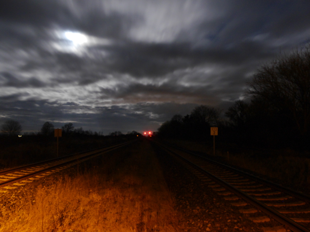 Eine zweigleisige Bahnstrecke bei Nacht, man sieht Wolken am Himmel und im Hintergrund zwei rote Signale.