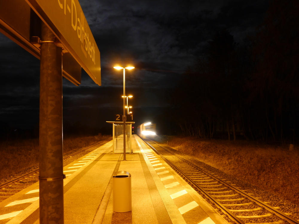 Ein hell erleuchteter Mittenbahnsteig bei Nacht, auf dem rechten Gleis fährt ein Regionalzug ein, der Bahnsteig ist menschenleer.