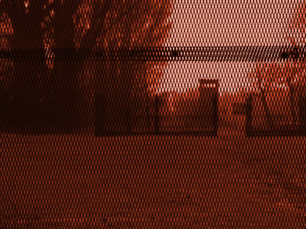 Ein bearbeitetes, rot eingefärbtes Foto. Durch das enge Gitter des Grenzzauns ist im Hintergrund der ehem. Grenzturm erkennbar.
