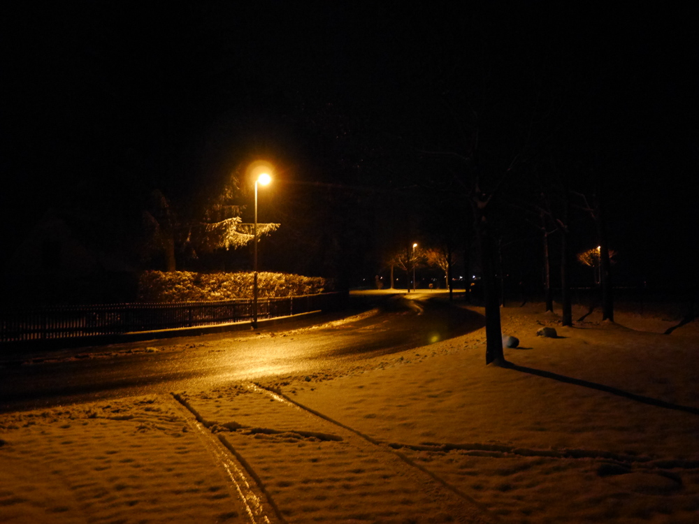 Eine nächtliche Straßenaufnahme an einer Kurve, es hat hat geschneit und nur die Straße ist gräumt. In der geräumten, Nassen Straße spiegeln sich die Lichter der Straßenlaternen
