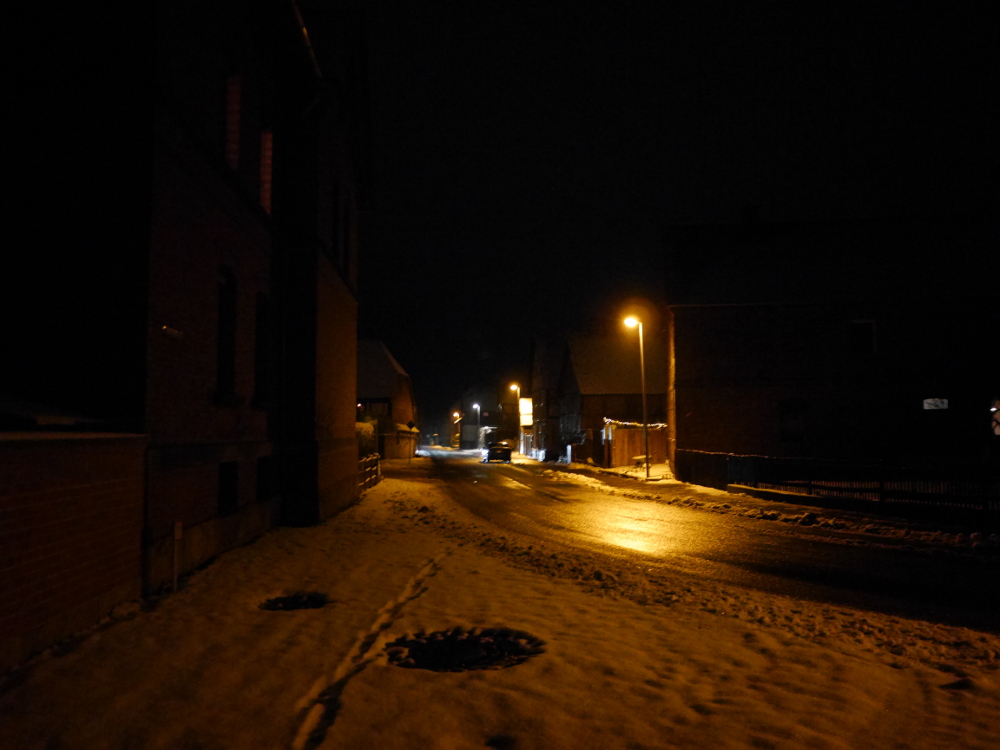 Eine nächtliche Straßenszene in einem Dorf, man sieht ein Auto auf der geräumten, nassen Straße stehen, im Verlauf der Straße wird das LIcht heller da von Links ein nicht-sichtbares Auto auf die Straße leuchtet.
