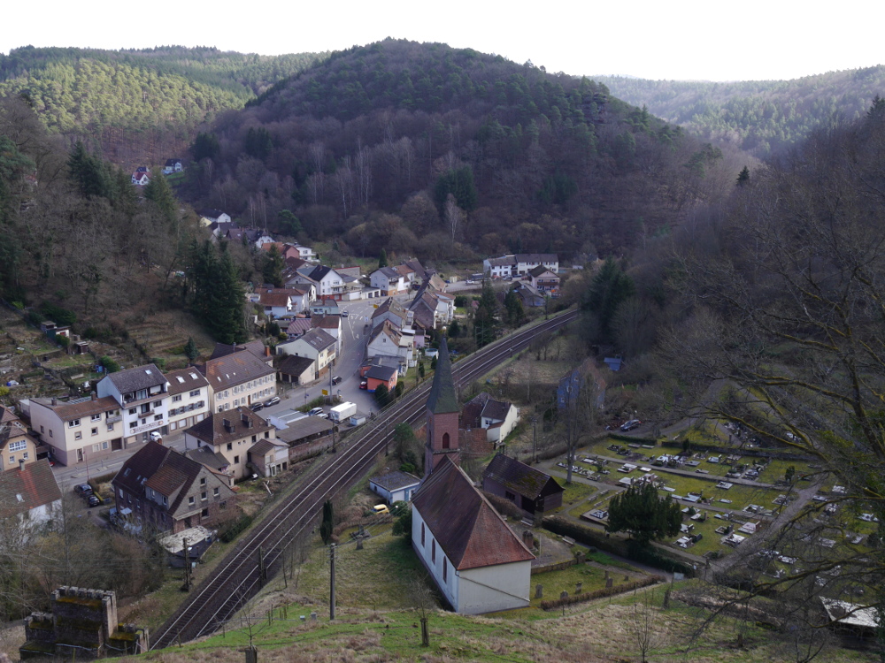Eine Landschaftsaufnahme der Stadt Frankenstein (Pfalz). Die Aufnahme ist von oben gemacht, man sieht die Bahnstrecke durchs Bild laufen, rechts davon eine Kirche mit Friedhof und Links die Häuser. Ziemlich in der Bildmitte ist auch ein bewaldeter Berg, an dem die Bahnstrecke nach rechts biegt. Links und rechts wird das Bild auch von bewaldeten Bergen begrenzt.
