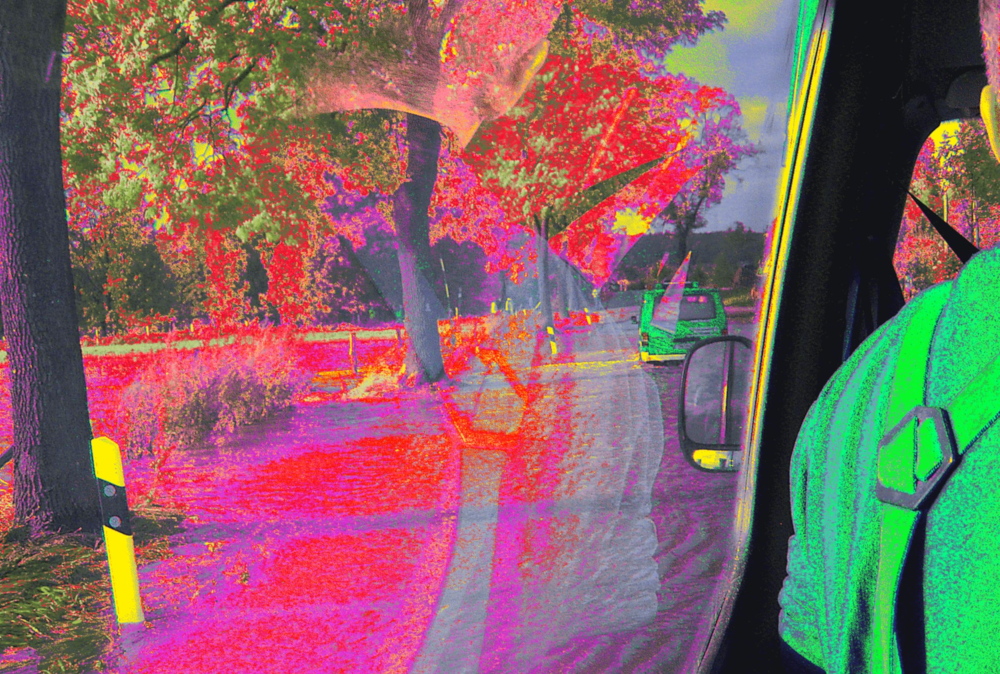 Das Bild ist stark farblich verfremdet, es zeigt den Blick aus einem Einsatzfahrzeug (Kleinbus), welches hinter einem anderen Kleinbus hinter her fährt. Auf der rechten Seite kann man den Rücken und die sich auf dem Rücken überkreuzenden Hosenträger eines Menschen erkennen. Beide Fahrzeuge fahren auf einer überfluteten Straßen, links der Straße stehen Bäume. Durch die Falrbveränderung sind die Bäume Rot, die eigentlich balen THW-Fahrzeuge grün und das Wasser fremdartig rot-lila.