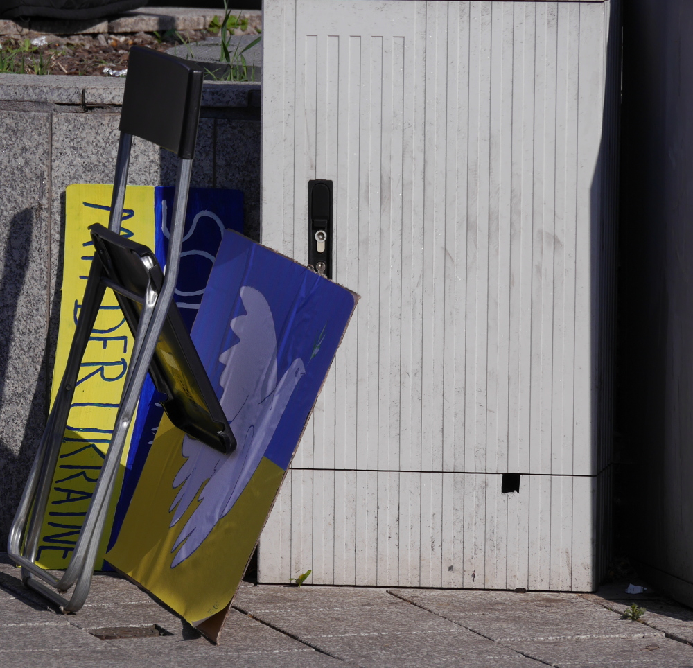 Das Foto zeigt am Rande einer Demonstration abgestellte Plakate und einen Klappstuhl. Eins der abgestellten Plakate ist die Fahne der Ukraine mit einer Friedenstaube, das andere zeigt den Schriftzug "Solidarität mit der Ukraine"