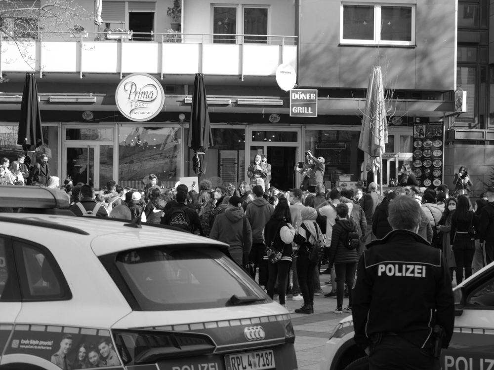 Ein s/w-Foto bei einer Demo in Ludwigshafen. Man sieht die Teilnehmer, die vor einer Frau steht, die eine Rede hält. Das Foto ist von etwas weiter hintern aufgenommen, man erkennt zwei Streifenwagen der Polizei und einen Polizeibeamten, der in Uniform dem Fotografen den Rücken zudeht.
