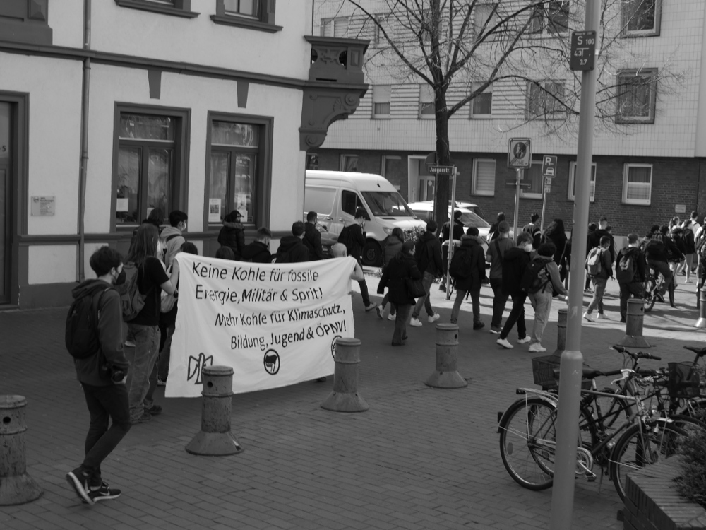 Das s/w-Foto zeigt Demonstranten und ein Transparent "Keine Kohle für fossile Energie, Militär & Sprit! Mehr Kohle für Klimaschutz, Bildung, Jugend & ÖPNV!".