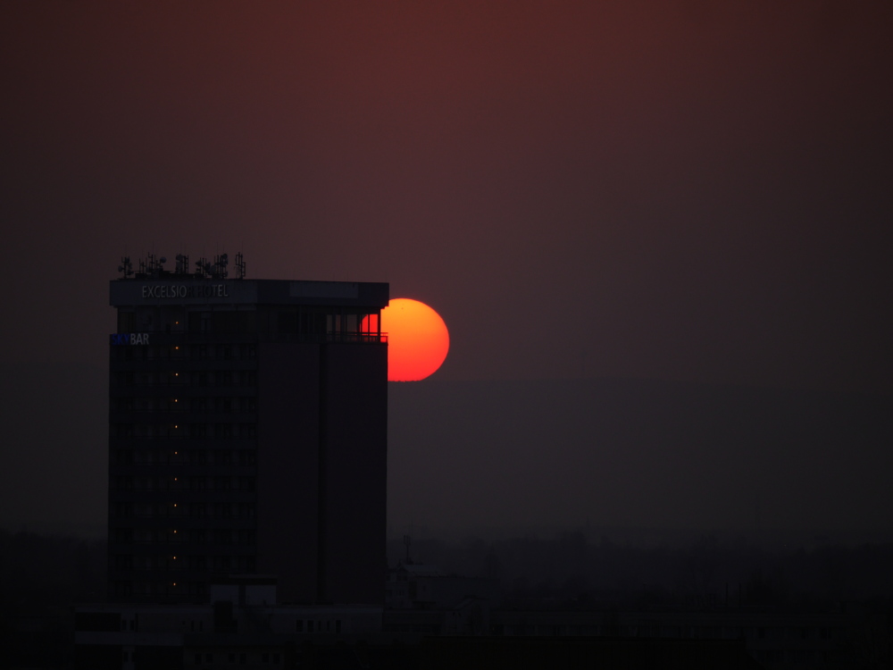 Das Foto zeigt einen Sonnenuntergang, die Sonne steht etwa auf der Höhe des oberen Stockwerks eines Hochhauses. Das Hochhaus hat ein Schild "Excelsiorhotel", wobei einige Buchstaben nicht mehr beleuchtet ist. Die Sonne ist gut als roter Feuerbal erkennbar und man kann auch sehen, das viel Staub in der Luft ist.