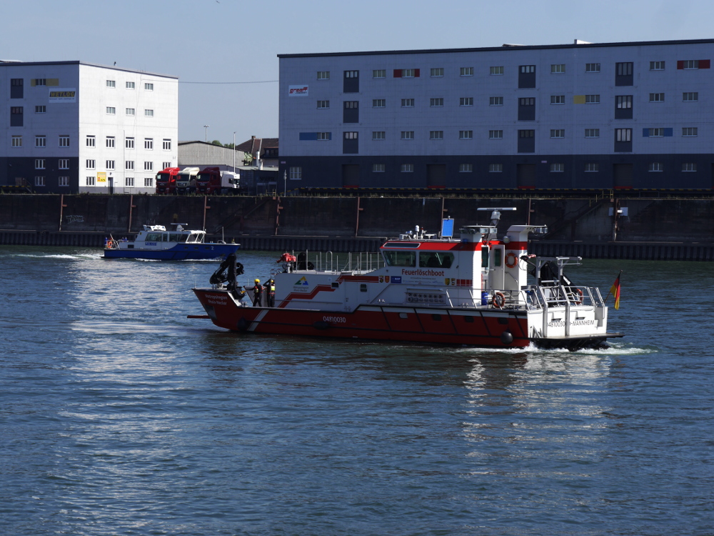 Ein großes Feuerlöschboot fährt auf dem Rhein, weiter hinten auf dem Rhein sieht man ein in Gegenrichtung fahrendes blaues Boot der Wasserschutzpolizei